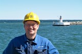 Man in blue jacket and yellow helmet in front of lighthouse